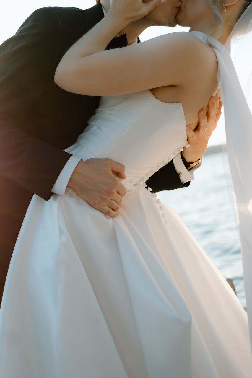 A bride and groom share a romantic embrace and kiss on their wedding day near a waterfront.