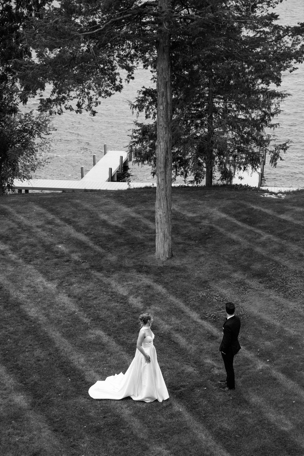 Black and white photo of bride and groom standing apart on lawn near lake with dock and trees.