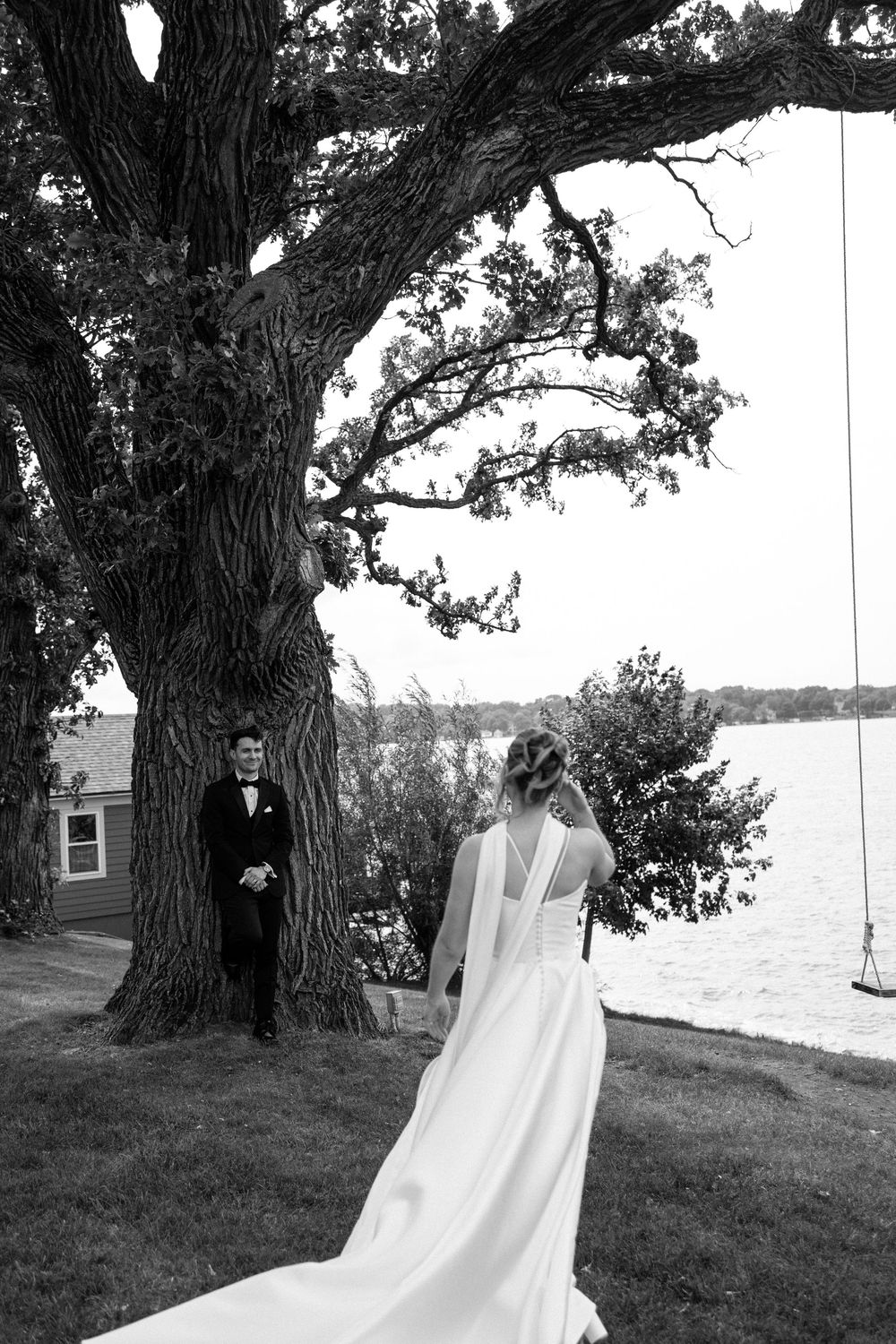 Black and white photo of a bride in a flowing gown approaching a groom in a tuxedo near a large tree by a lake.