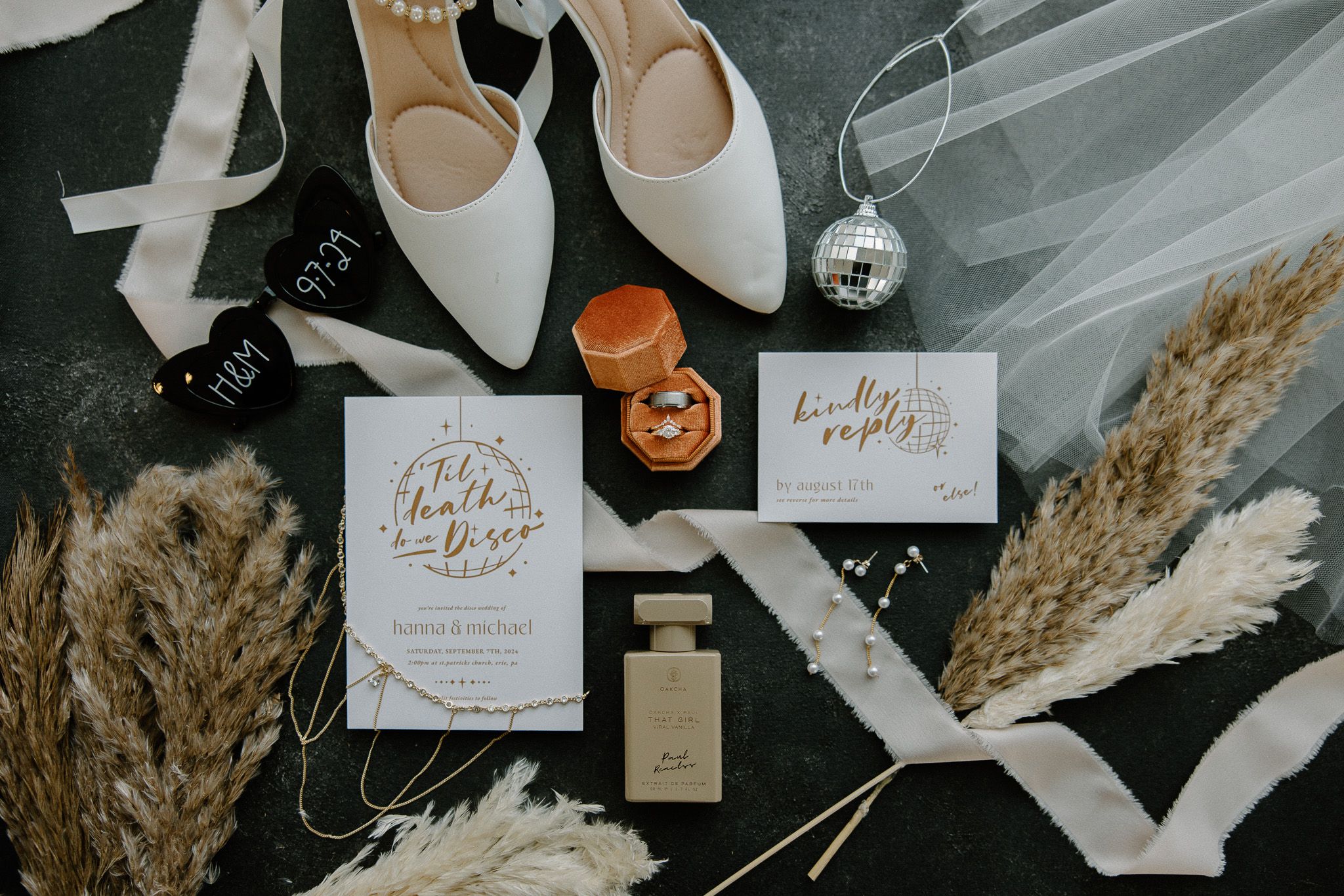 Wedding details flatlay with white shoes, invitations, dried pampas grass, and golden accents on dark background.