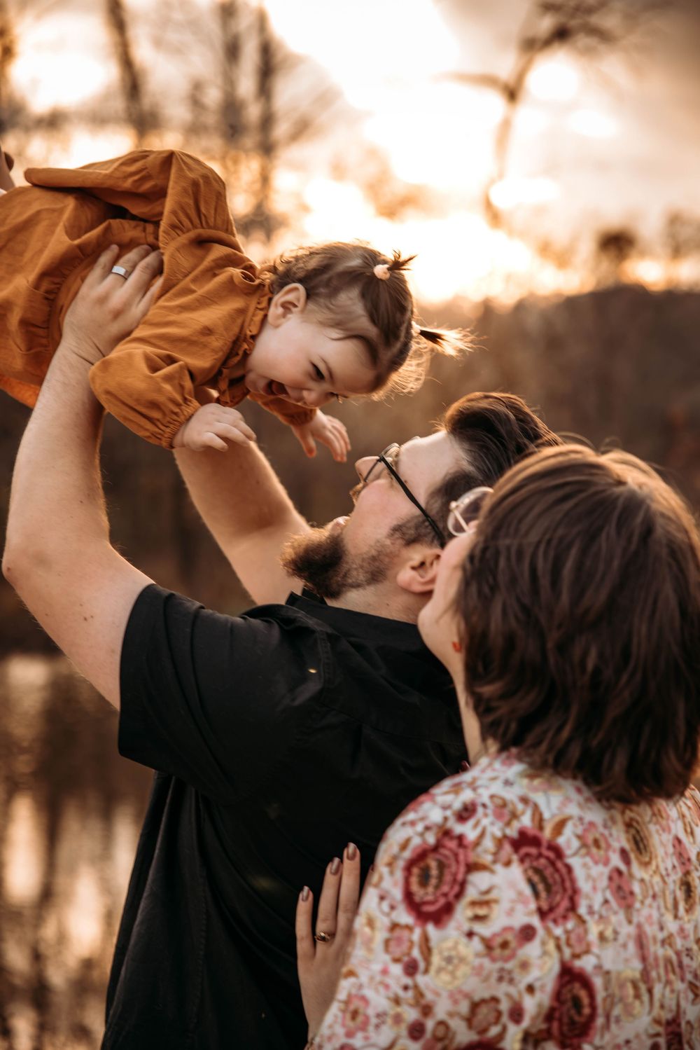 Emotional motherhood photo session featuring a father lifting his baby in the air in a wooded Branson, Missouri setting.