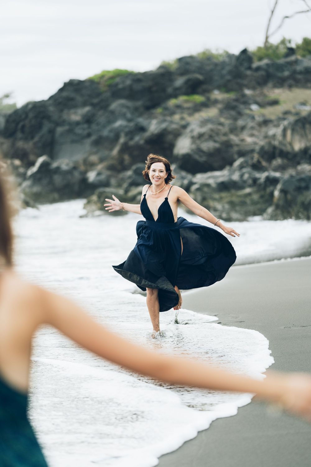 A person in a flowing black dress dances gracefully along a scenic beach shoreline with waves rolling in.