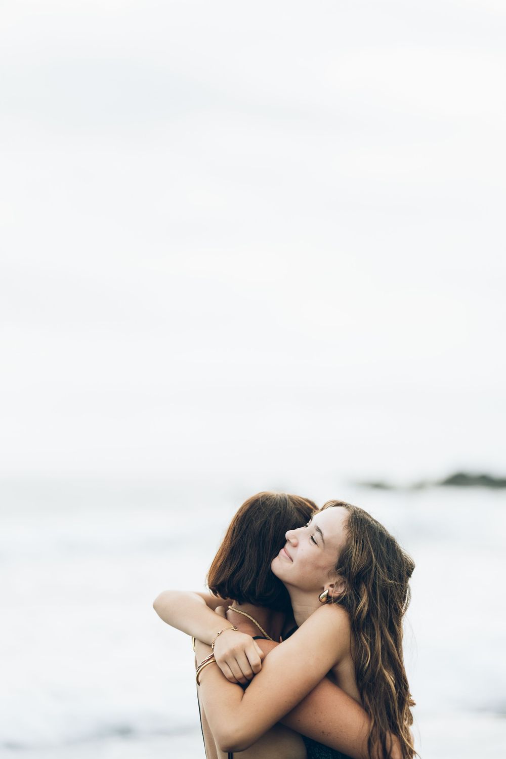 Two friends share an emotional embrace on a misty beach with the ocean in the background.