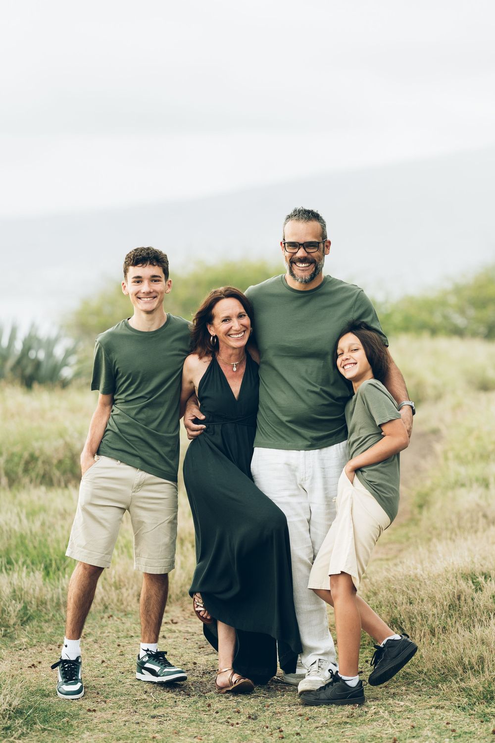 Happy family of four posing outdoors in coordinating green and neutral outfits on a grassy hillside.