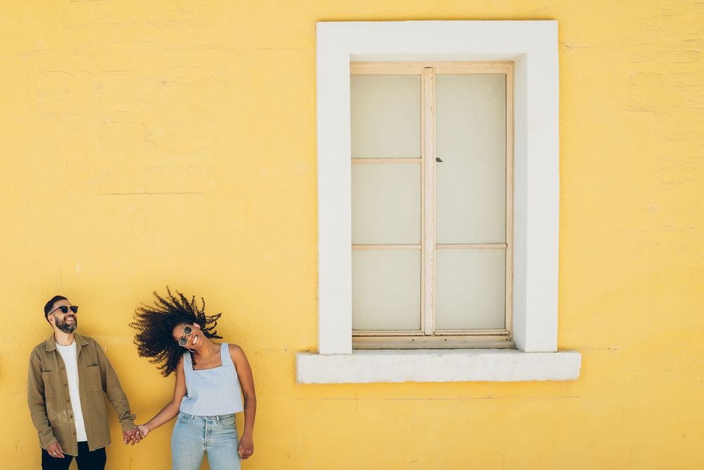 Couple holding hands against a bright yellow wall with a white window, woman's hair flying in the wind.