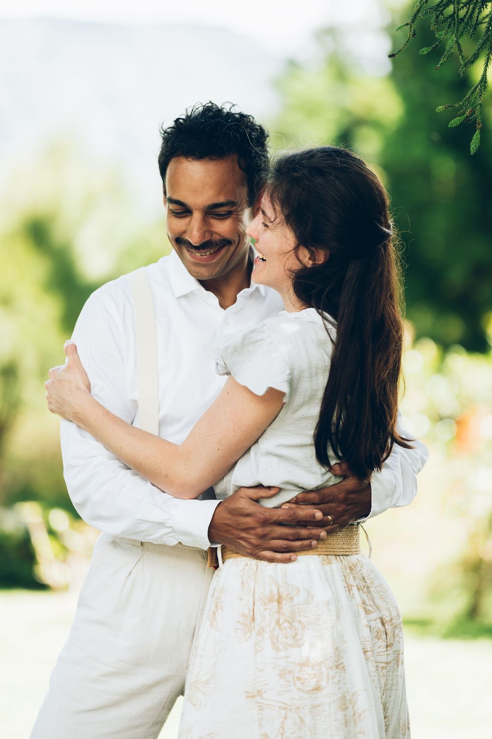 A couple in white outfits embraces joyfully outdoors in a garden setting with soft natural lighting.