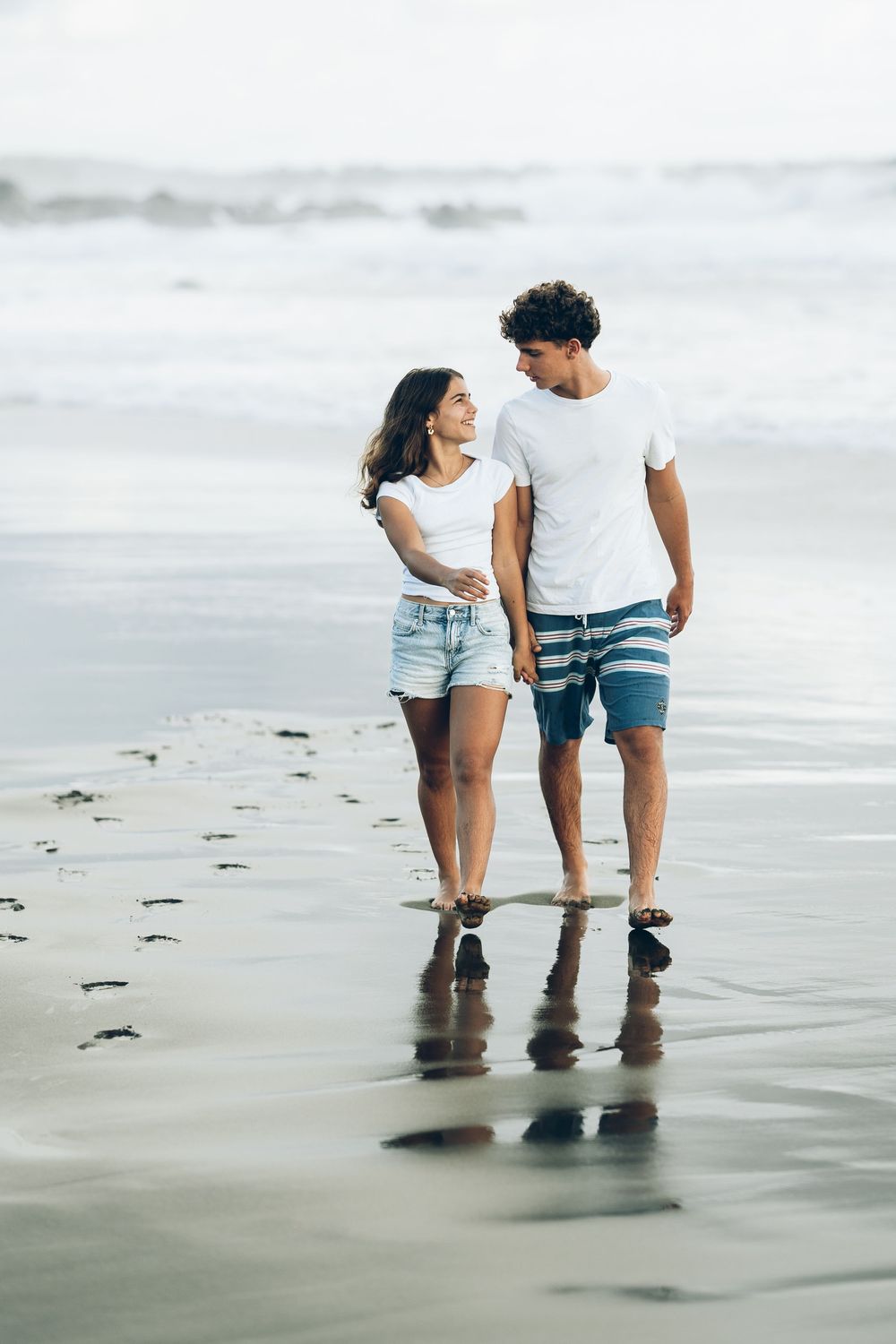 A young couple walks hand in hand along a wet sandy beach, smiling at each other, wearing casual summer clothes.