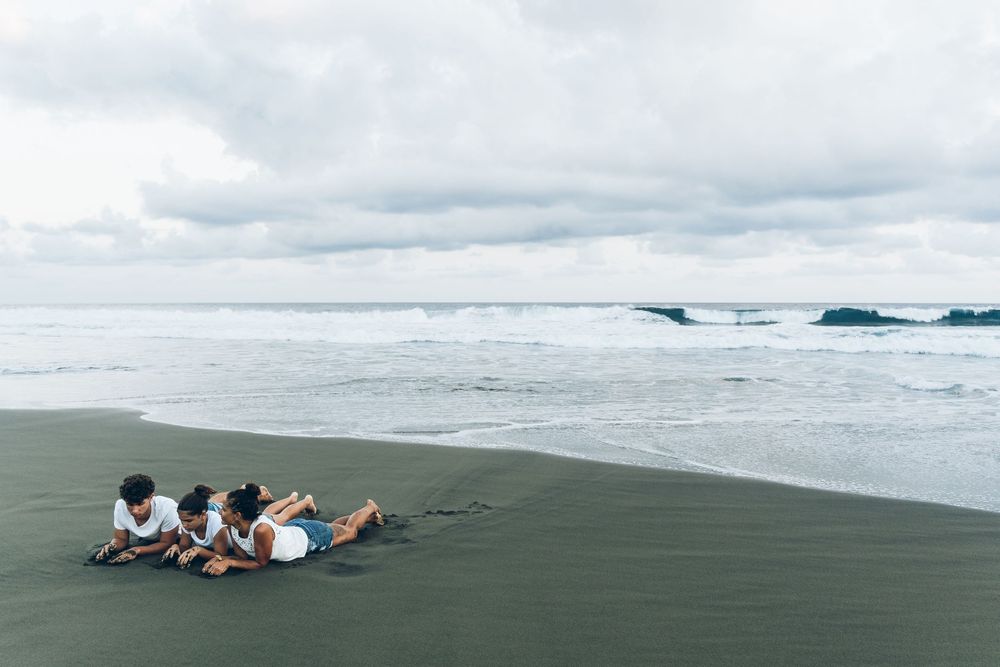 Three people lying on dark sandy beach shore watching ocean waves under cloudy sky.