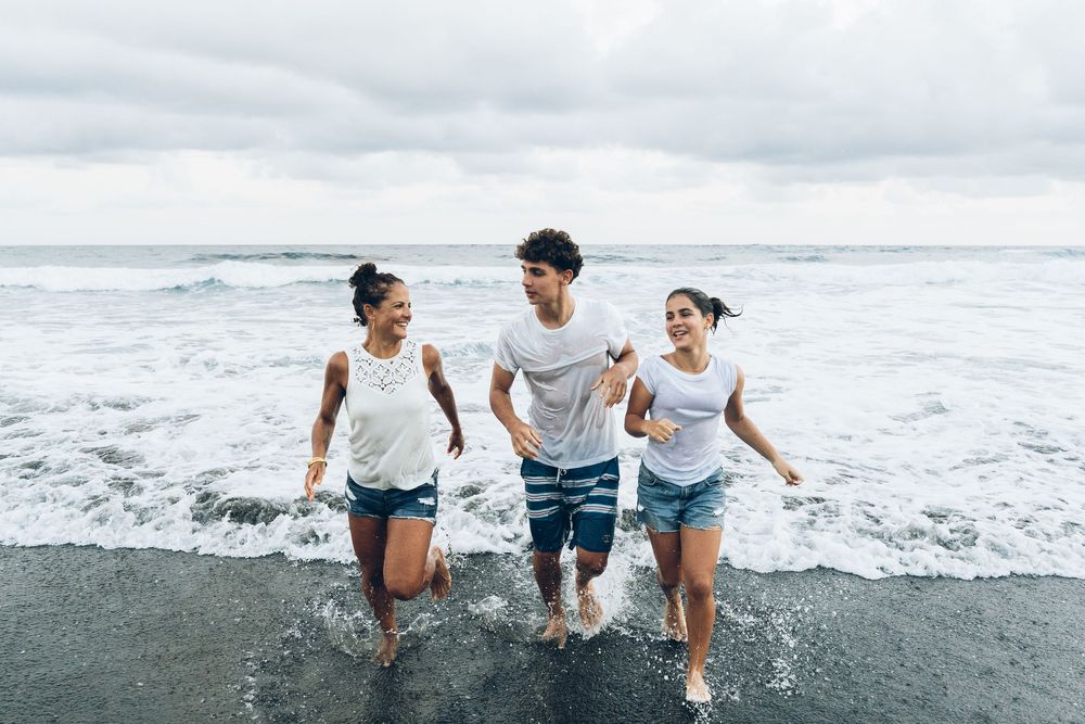 Three friends running joyfully through ocean waves on a black sand beach on a cloudy day.
