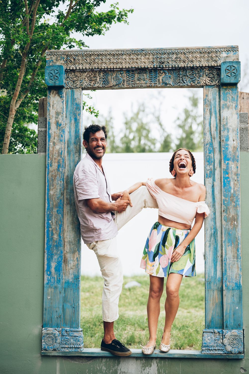 A couple shares joyful laughter while posing playfully in a rustic blue-framed window outdoors.