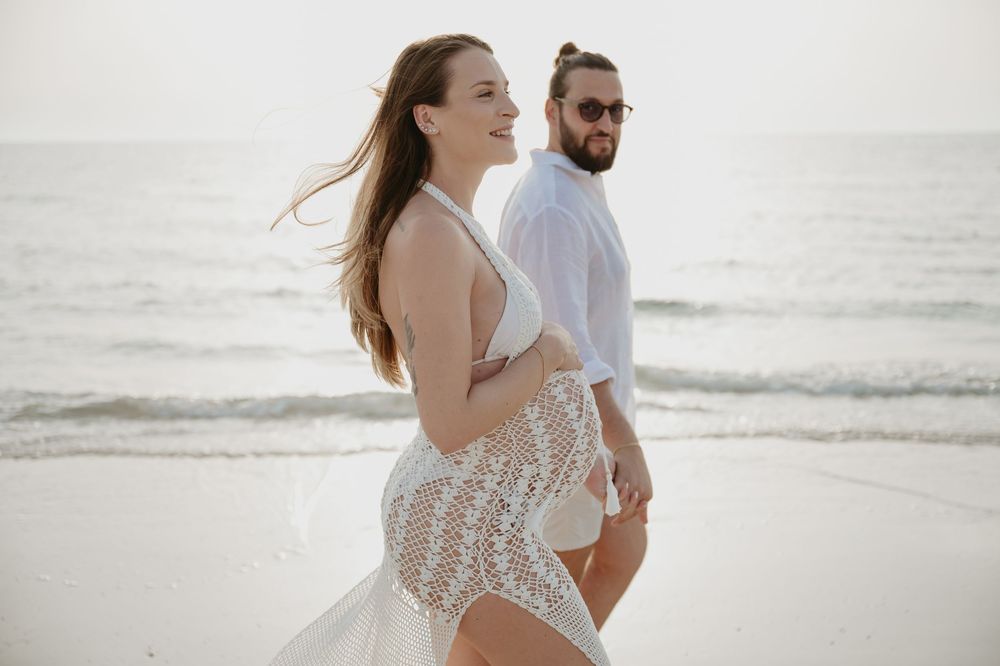 Couple in white outfits walking on a beach, woman in lace dress smiling, man in white shirt with sunglasses.