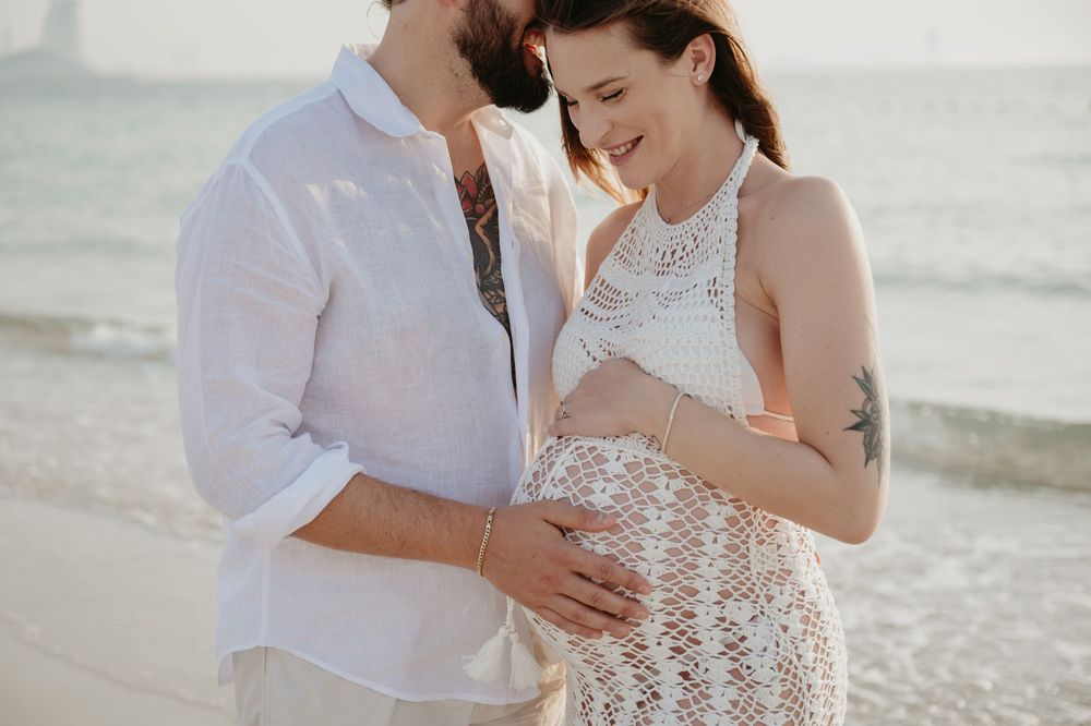 Couple tenderly cradling pregnant belly on a sunny beach, both dressed in white, sharing a loving moment.