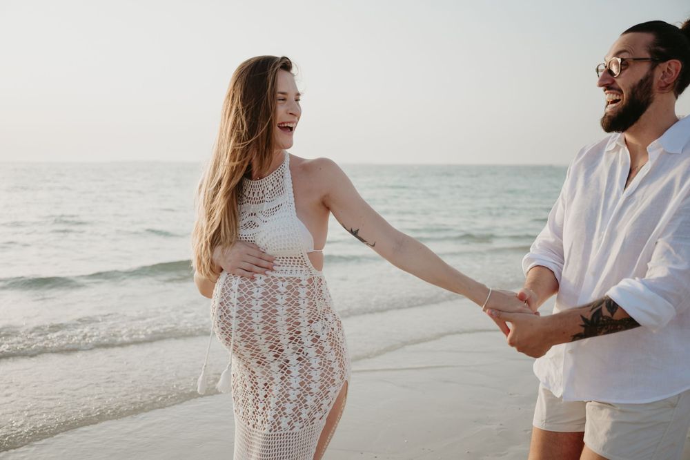 Happy couple holding hands walking on a beach, woman in white lace dress, man in white shirt, ocean background.