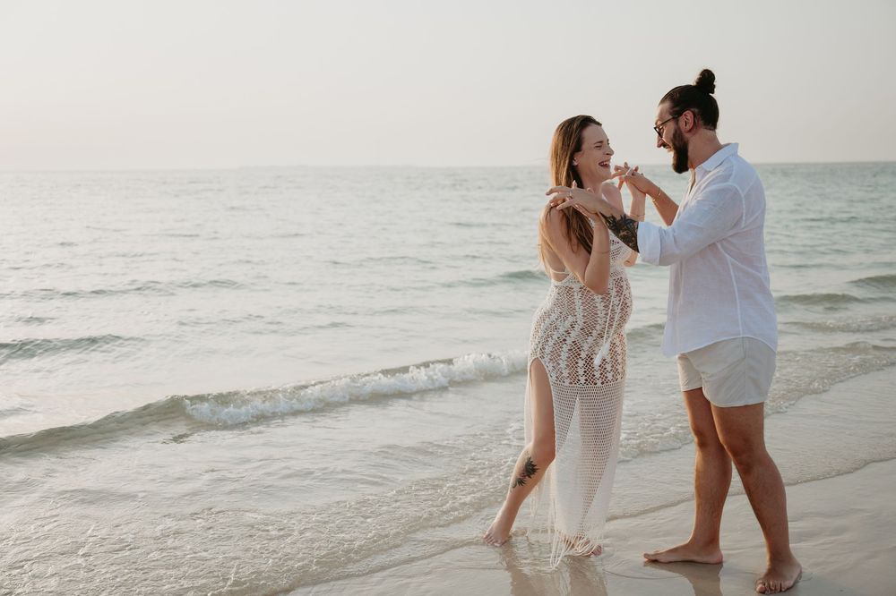Couple dancing romantically on a sandy beach at sunset, woman in white lace dress, man in white shirt.