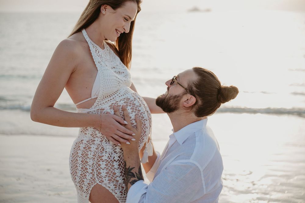 Pregnant woman in lace dress with partner kneeling on beach at sunset, touching her baby bump.