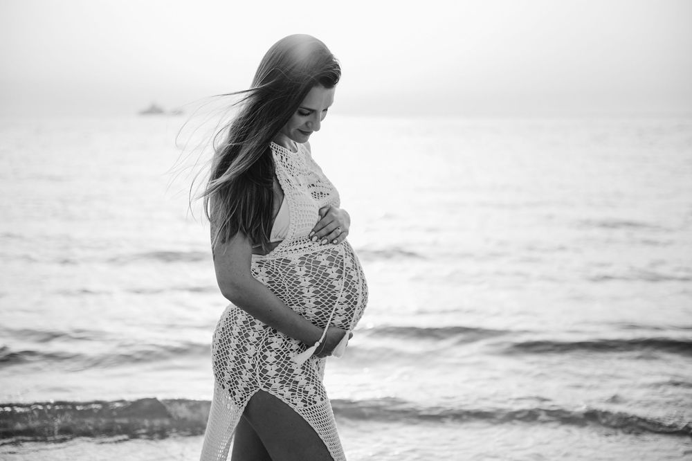 Black and white photo of a pregnant woman cradling her belly on a beach with ocean waves in the background.