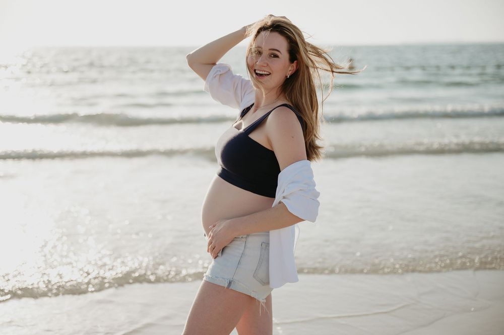 Smiling pregnant woman in black crop top and shorts poses on sunny beach with ocean waves in background.