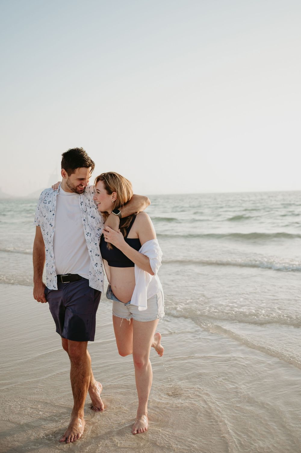 Couple walking barefoot on a sandy beach, embracing and smiling at each other with ocean waves in the background.