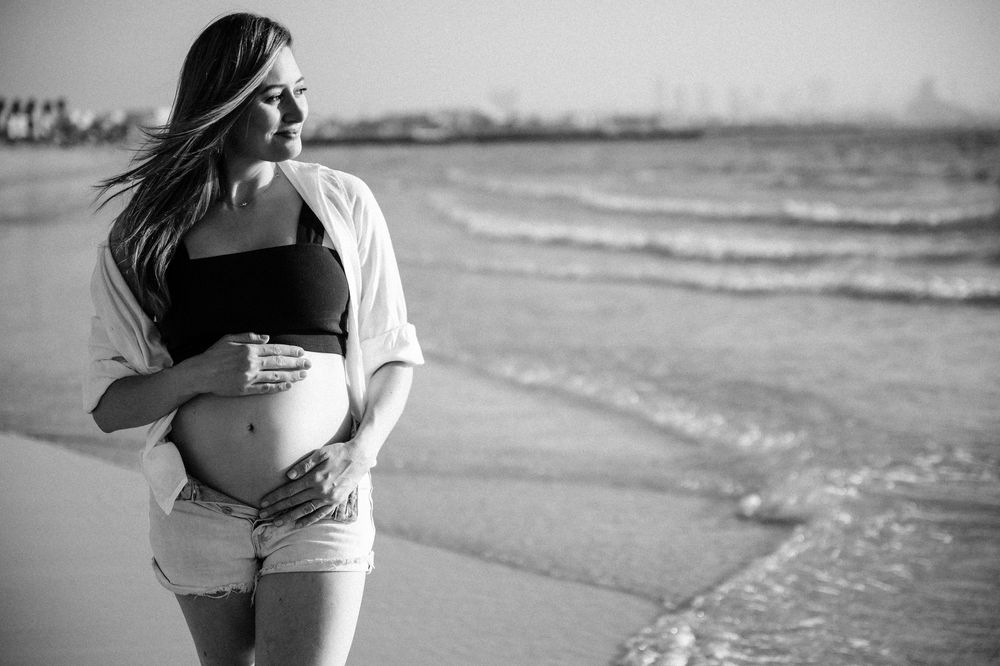 Pregnant woman in black crop top and shorts cradling her baby bump on a sunny beach in black and white.