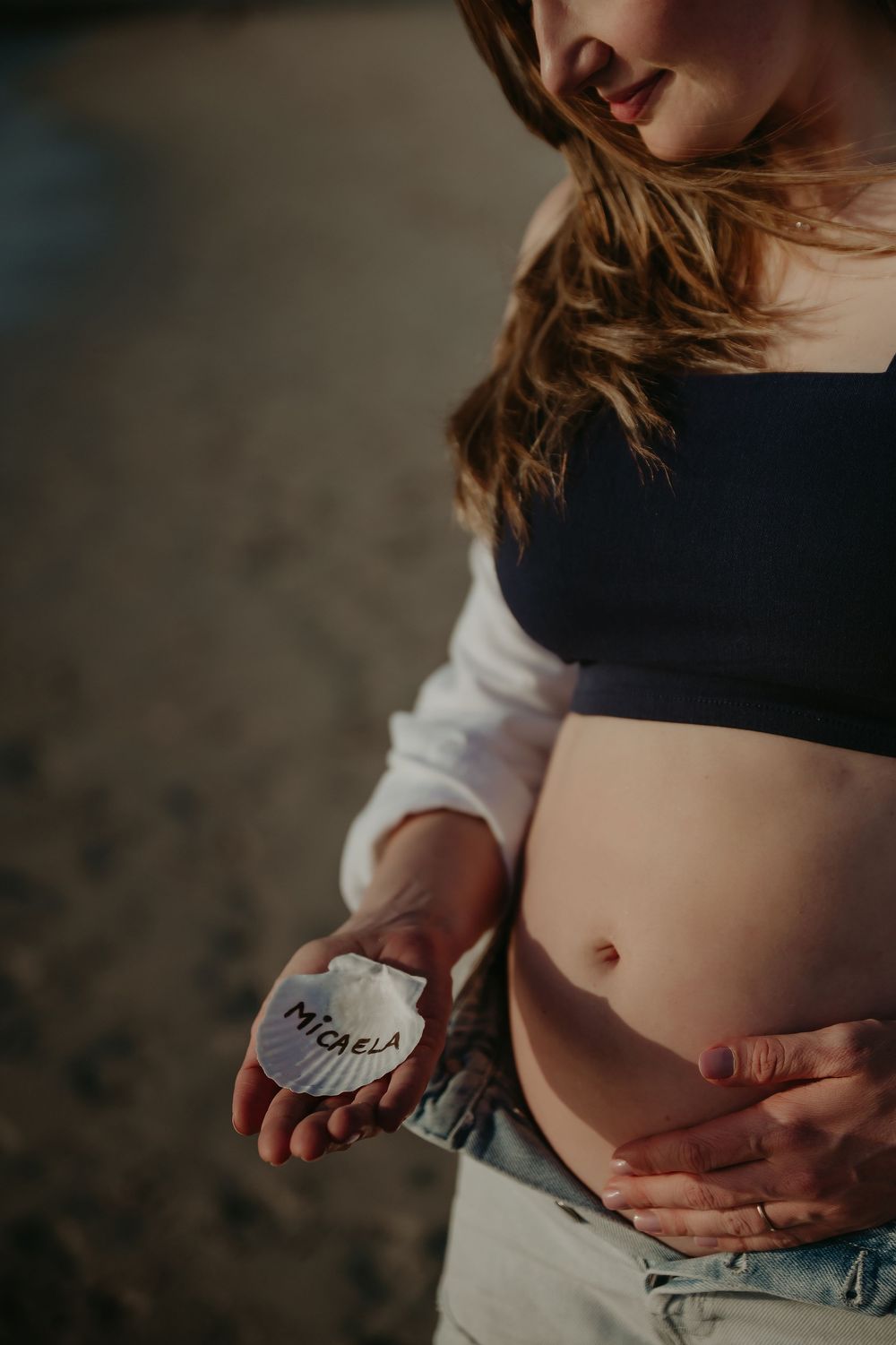 Pregnant woman holding a small stone engraved with the name Micaela, cradling her baby bump outdoors.