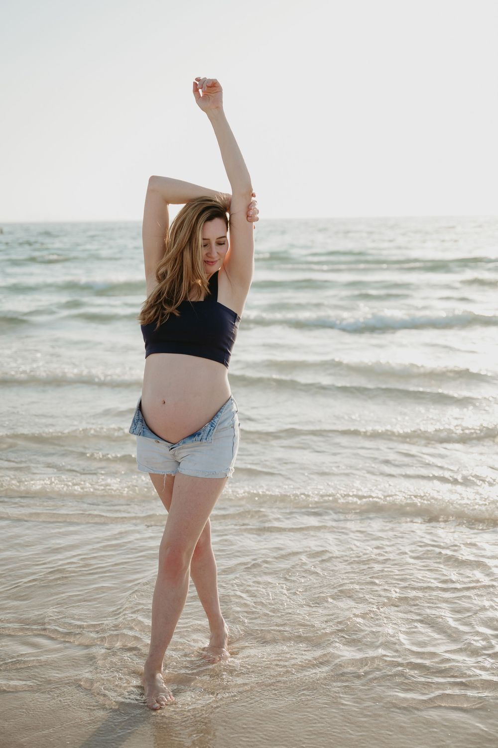 Pregnant woman in black crop top and denim shorts posing on beach with arms raised, ocean waves in background.