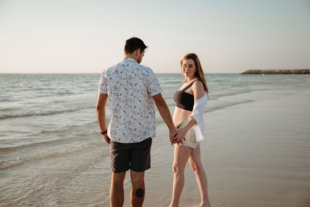 Couple holding hands on a sandy beach at sunset, woman in black bikini top and white shorts.