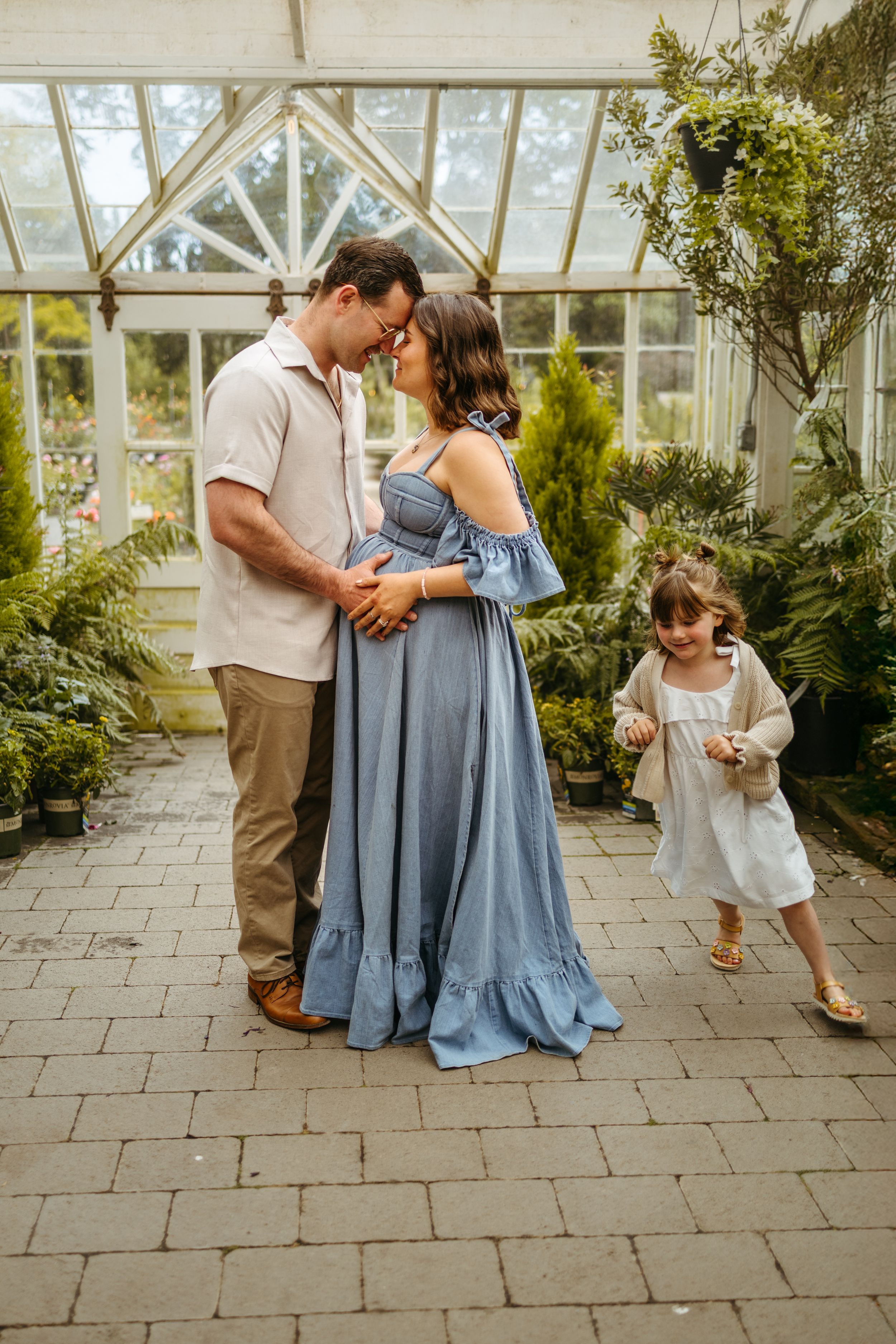 A romantic moment between a couple in a greenhouse with a young child wearing white running past on stone pavement.