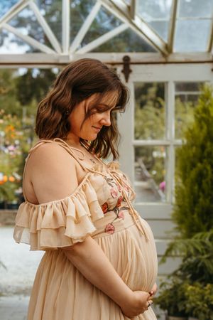 An elegant maternity photo in a greenhouse features a flowing peach-colored dress with ruffled sleeves.