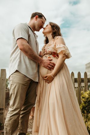 Romantic maternity photo showing couple in casual attire embracing by rustic wooden fence under cloudy sky.