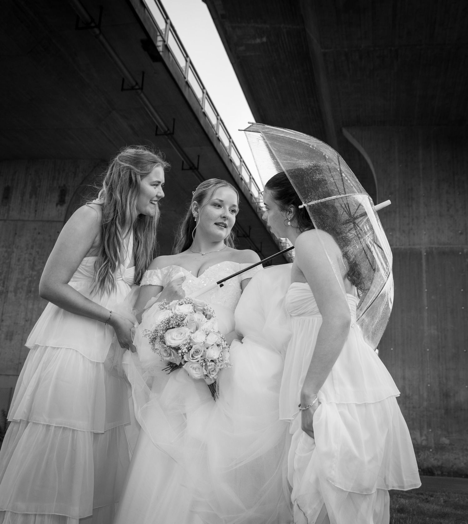 Bride and two bridesmaids in white dresses share a joyful moment under a clear umbrella.