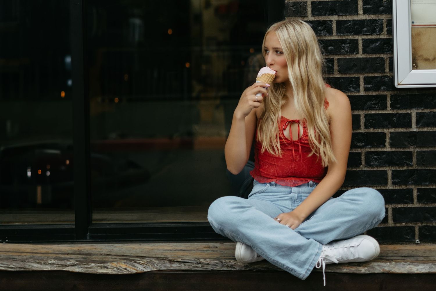 Someone in a red tank top and light wash jeans sits cross-legged on a wooden bench while drinking from a cup.