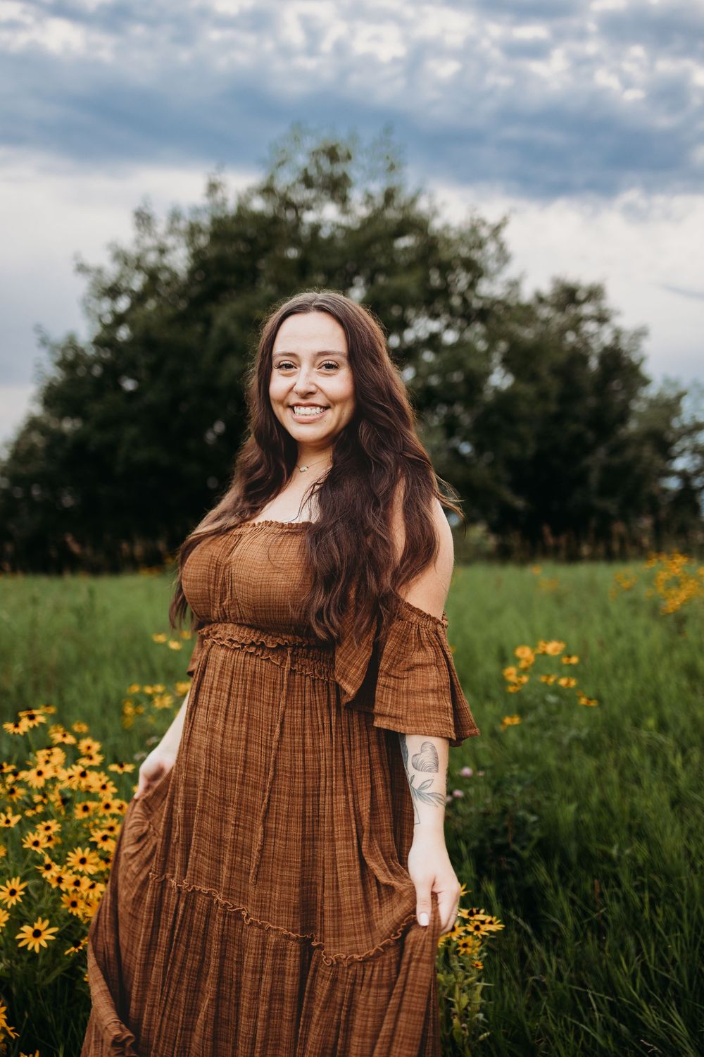 A brown off-shoulder prairie dress stands out against a field of yellow wildflowers on a cloudy summer day.
