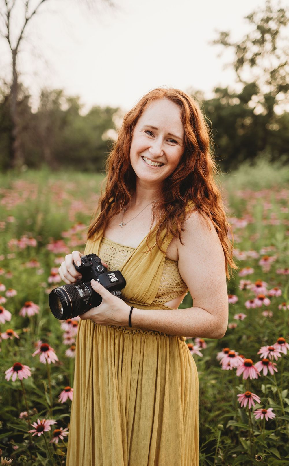 A photographer in a yellow dress holds a camera while standing in a field of pink coneflowers at sunset.