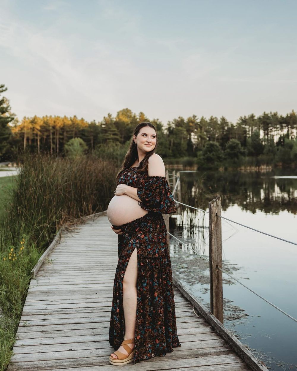 A tranquil wooden dock extends over a calm lake surrounded by pine trees at sunset with a floral dress visible.