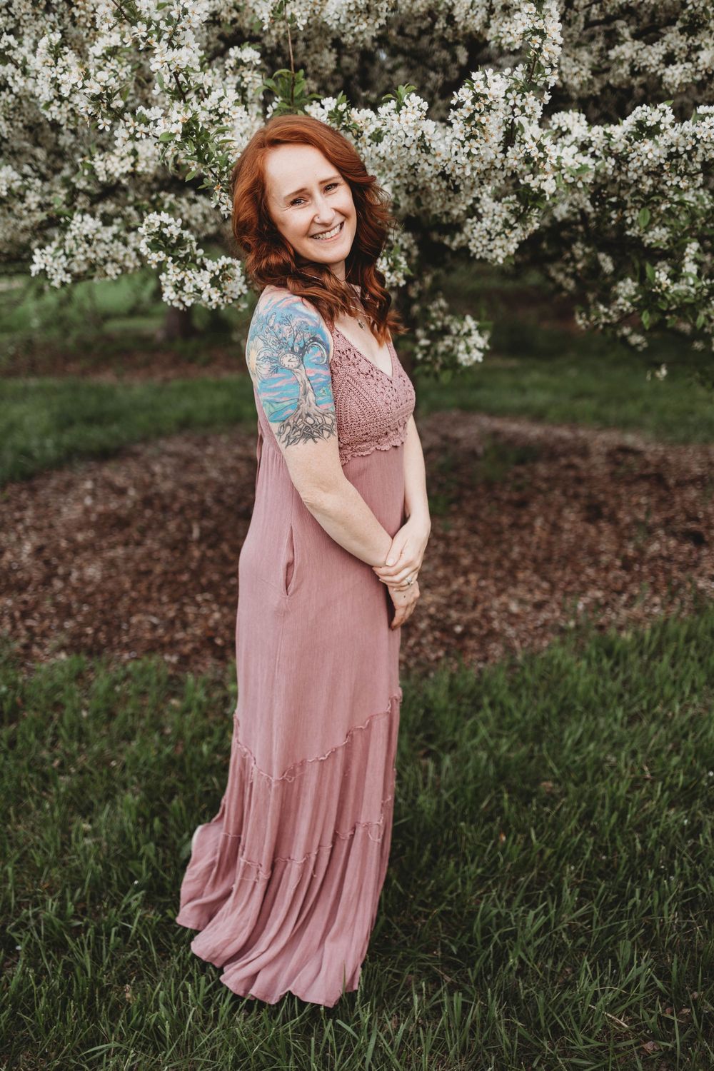 A woman in a flowing dusty pink maxi dress poses outdoors near blooming white spring flowers and grass.