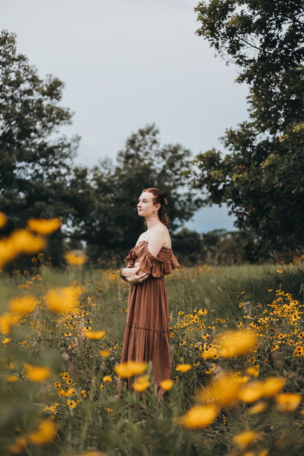 Woman in vintage brown dress stands in field of yellow wildflowers during golden hour surrounded by trees.