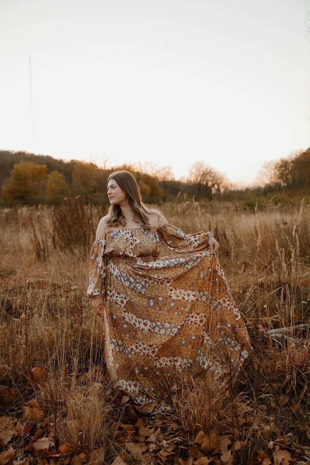 Woman in floral boho maxi dress standing in autumn field at golden hour sunset.