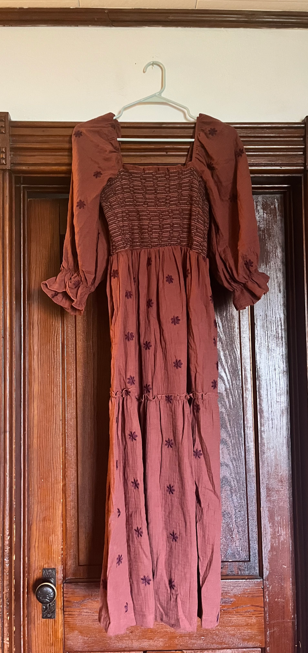 Rust-colored maxi dress with smocked bodice, puff sleeves, floral embroidery, and tiered skirt on wooden hanger.