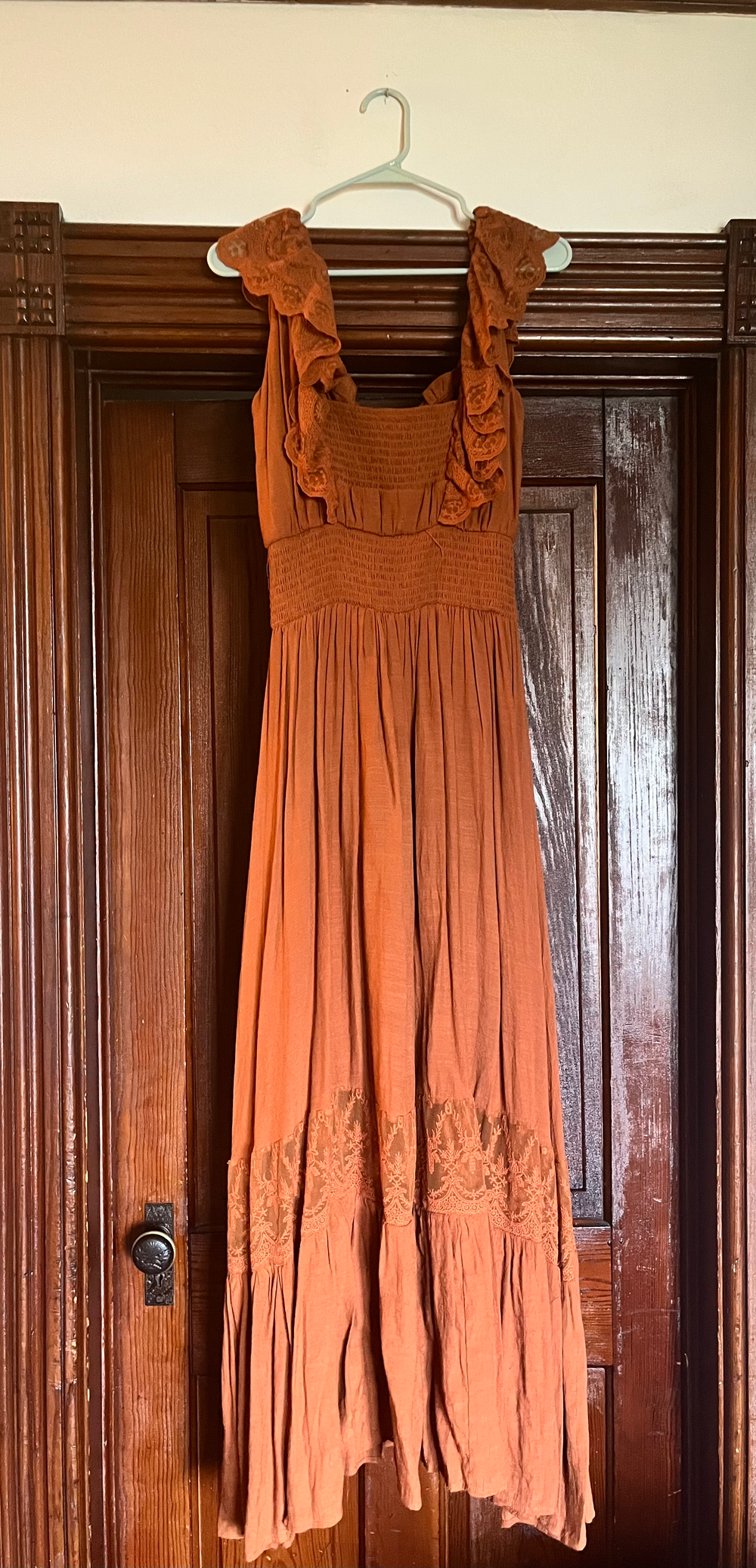 Rust orange maxi dress with smocked bodice, ruffle straps, and embroidered hem detail, hanging on wooden door.