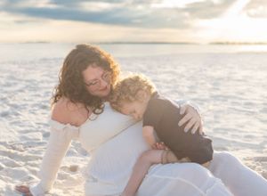 A little boy snuggles pregnant mother on the sand with sun shining through clouds Pensacola, FL