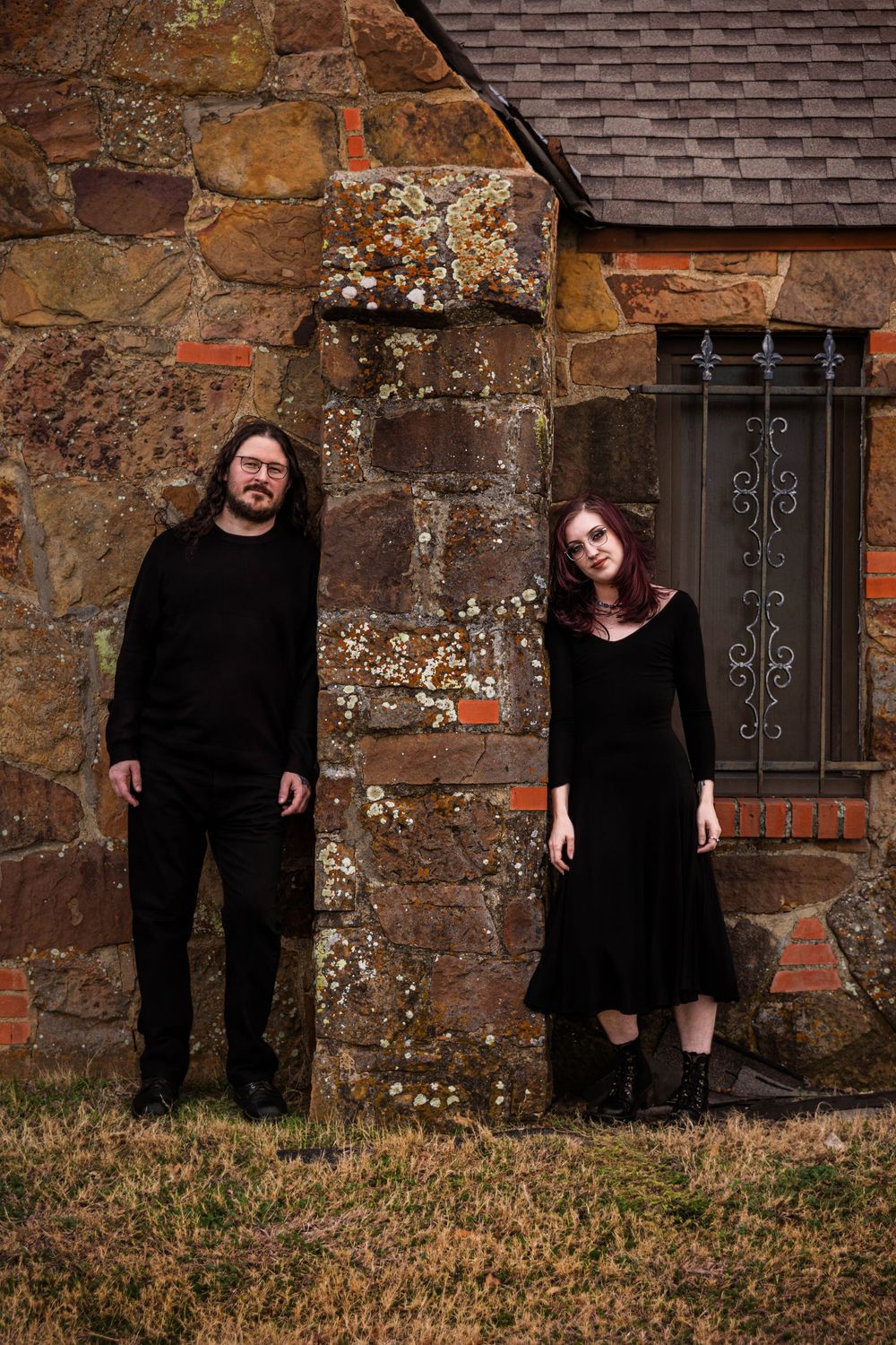 A couple poses in front of a rock building. They are wearing all black. This photoshoot was done in a cemetery.