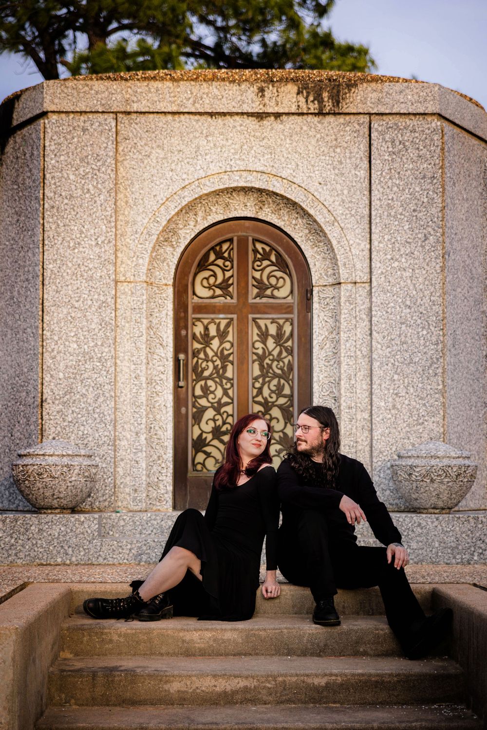 A couple poses in front of a beautiful mausoleum in a cemetery.