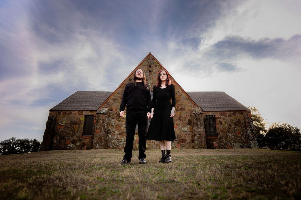 A couple poses in front of a rock building. They are wearing all black. This photoshoot was done in a cemetery.