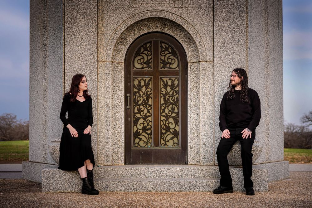 A couple poses in front of a beautiful mausoleum in a cemetery.
