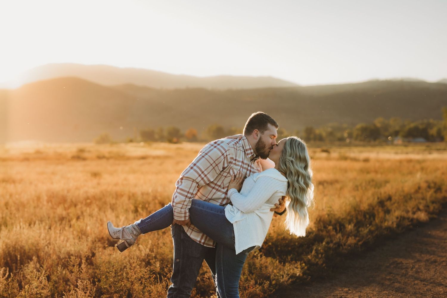 Romantic couple embraces in golden field during sunset with mountains in background.