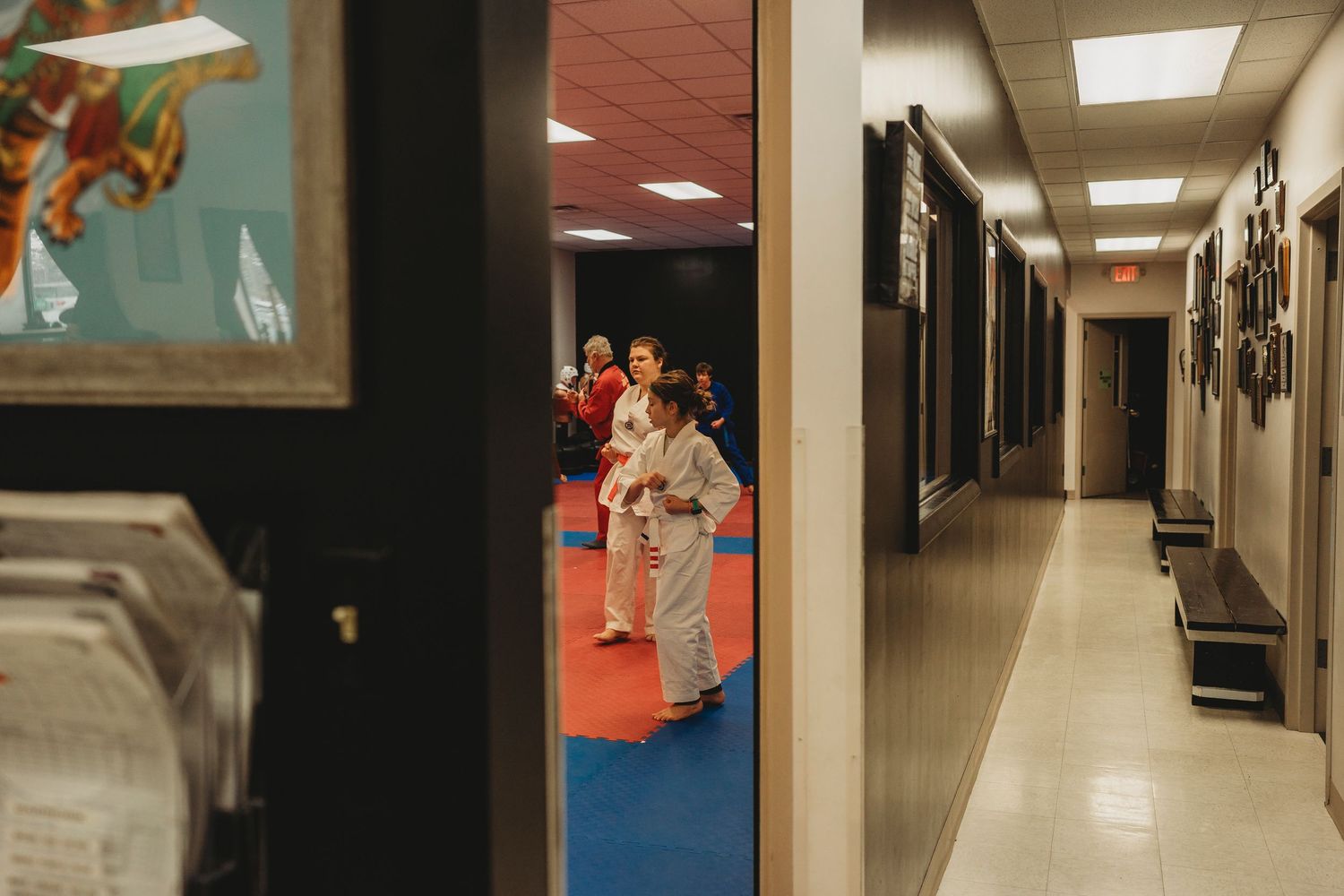 A long institutional hallway with fluorescent lighting and tiled floors leads into darker areas with exit signs visible.