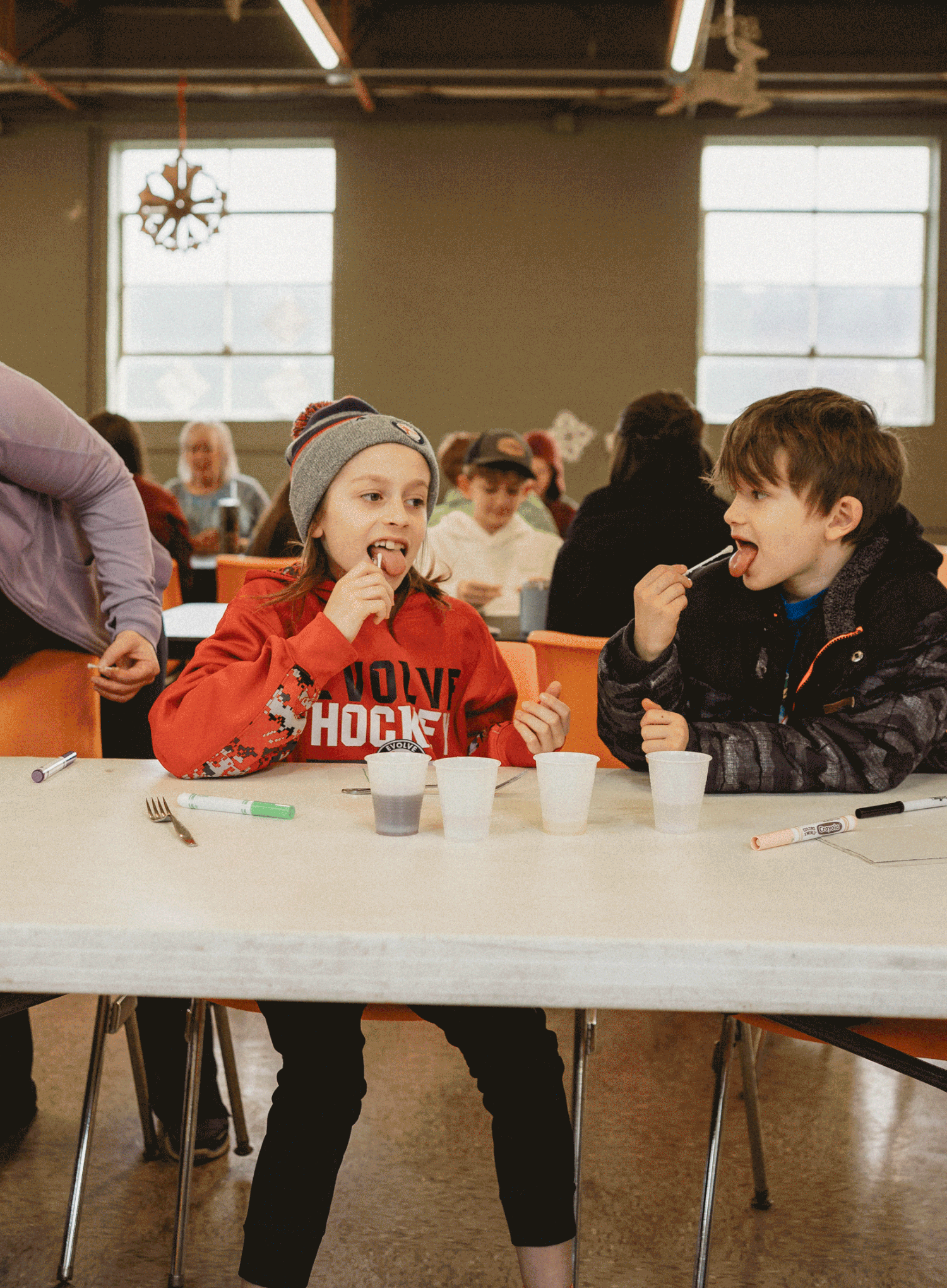 Two students enjoy a snack break while sitting at a long white table in a cafeteria setting.