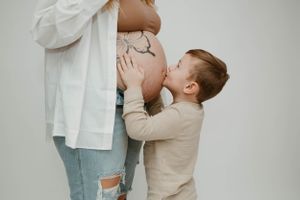 Young child kisses pregnant belly while standing against light grey studio background.
