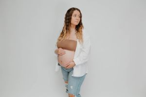 Maternity portrait in white shirt and distressed jeans against studio backdrop.