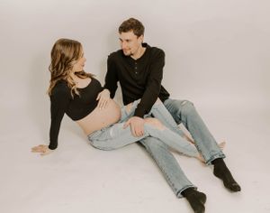 A couple in black tops and jeans sit casually together against white backdrop.